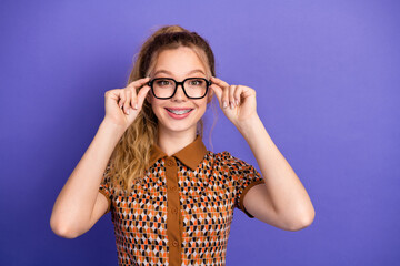 Smiling young woman with glasses adjusts her frames in a geometric patterned dress against a purple...
