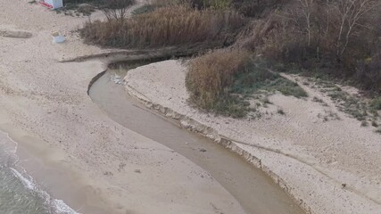 Dynamic aerial drone view of a narrow coastal river bend flowing between dense autumn bushes and sandy shoreline on a windy seaside landscape - Powered by Adobe