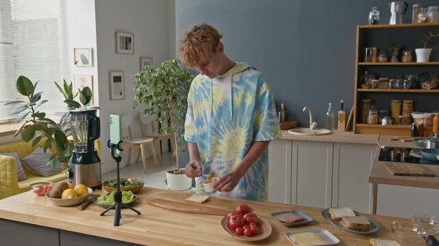 Gen Z Caucasian boy standing at kitchen counter, making sandwich and recording video for blog via smartphone on tripod