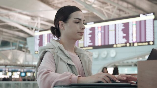 Young woman with dark hair, focused on laptop in modern airport terminal, surrounded by travelers, showcasing digital connectivity and travel experience