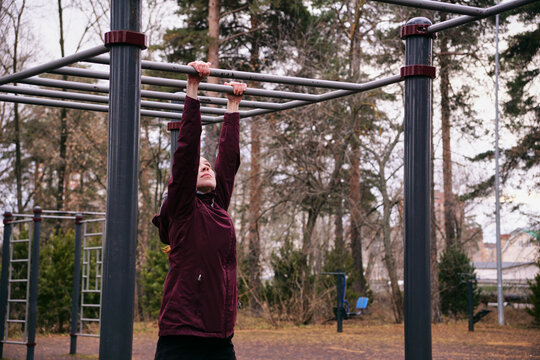Young sports woman hanging on horizontal bar during workout on fresh air. Active brunette in sport outfit training abdominal muscles at outdoor sport