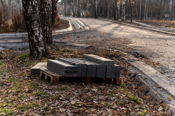 Stack of gray concrete paving stones on a wooden pallet next to a partially completed park path renovation in an autumn setting.