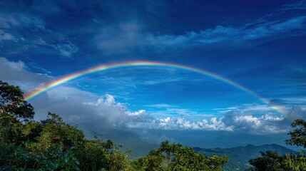 Obraz premium A vibrant rainbow stretches across the sky connecting lush green mountains and bright clouds after rain.