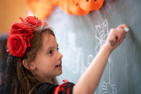 Girl with a flower wreath on her head wearing a black sweatshirt drawing a spider web with chalk on a blackboard. Creative Halloween classroom activity in Germany.