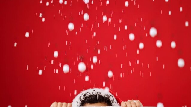 Cheerful young boy peeks over a white surface wearing a Santa hat against a bright red background, radiating playful holiday joy and festive excitement.