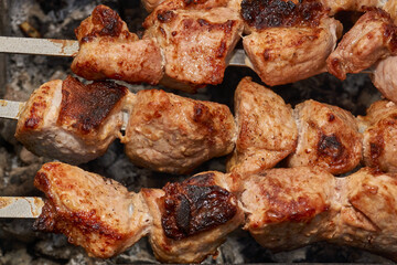 Large pieces of pork being grilled on skewers over hot charcoal. Delicious barbecue with a golden crust cooking outdoors during a picnic or countryside gathering.