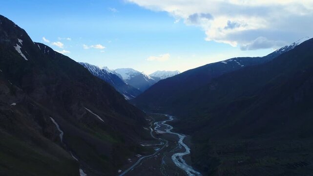 Aerial view of a winding river carving through a deep valley, flanked by towering mountains partially covered in snow, creating a stunning contrast, Naran, Khyber Pakhtunkhwa, Pakistan.