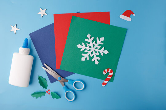 Colored paper sheets with cutout snowflake, scissors, glue bottle and Christmas paper elements on blue background. Flat lay composition with copy space. Winter holiday and Christmas craft concept,