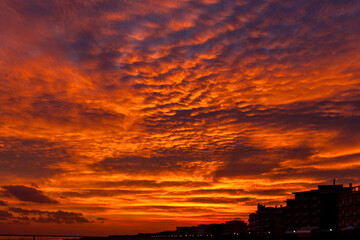 Dramatic sunset sky ablaze with orange and purple hues above silhouetted coastal buildings. A vivid display of nature’s evening light.