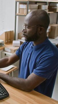 Medium vertical arc shot of African American male delivery company operator working on computer in office, preparing shipping documents, female colleague scanning barcodes near packages on shelves