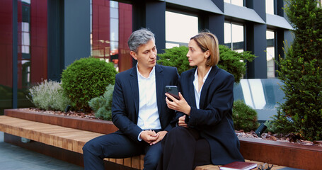 Business colleagues man and woman sitting and discussing work using smartphone outside office. City meeting and phone with business people outdoor on rooftop for collaboration or planning