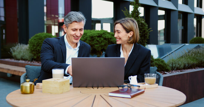 Business couple smiling and interacting while using laptop sitting on the summer terrace of the cafe . Two business people man and woman working with modern devices drinking coffee sitting on the