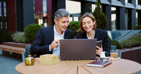 Business partners coworkers businessman and businesswoman working together in city with laptop writing notes in notebook. Man and woman in suits having fun, laughing, watching something funny on