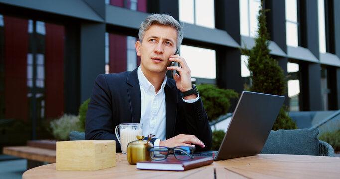 Middle-aged businessman working on laptop at an outdoor cafe while having a business call on phone. Focused mature man having online meeting sitting on terrace near office building,