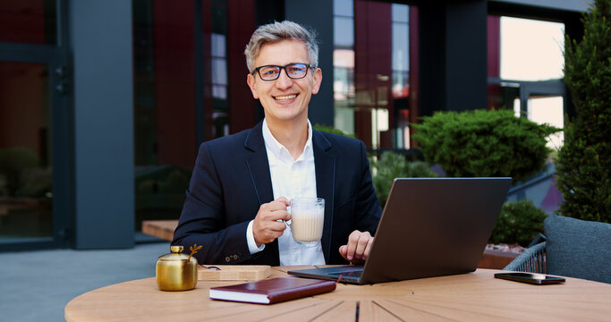 Close up portrait smiling face businessman wearing stylish glasses looking at camera sipping coffee and typing on laptop posing outdoors during lunch break of the cafe. Male manager employer