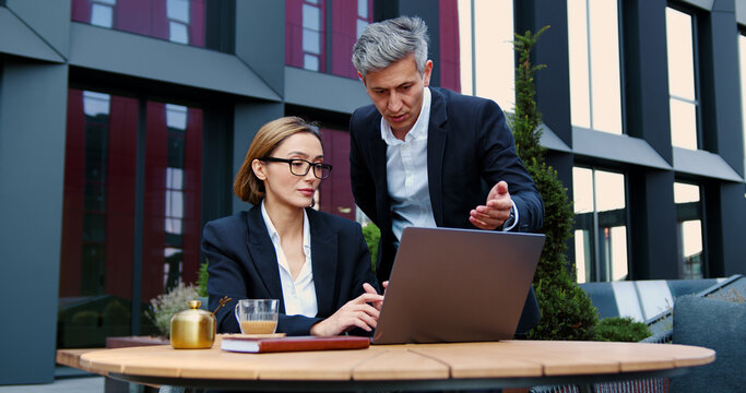 Professional man woman using laptop discussing project sitting in cafe outdoors. Attractive business lady with computer device listening to businessman. Both smiling and interacting in urban sunny