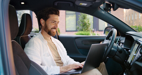 Handsome Caucasian man working remotely on a laptop while sitting in his parked car near a modern office building, focused on his task