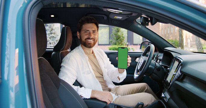 Confident young businessman sitting inside a car, smiling and pointing at a smartphone with green screen. Mobile phone mockup with chroma key for app or website presentation.