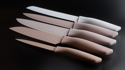 Set of five modern pastel beige and white kitchen knives arranged diagonally on a dark black background. Professional chef cutlery with silicone handles and colored blades. Photo