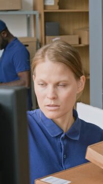 Vertical shot of focused Caucasian female e-commerce delivery company operator, holding heap of parcels, working on computer to process new orders, black male colleague filling report on tablet