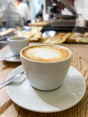 Coffee cup with latte art on wooden table, accompanied by a small espresso cup, showcasing a cozy cafe atmosphere with blurred background elements and inviting ambiance