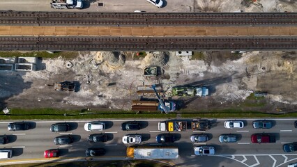 Aerial view of road construction repair site with heavy machinery, workers, next to moving car traffic and railway system on a sunny day