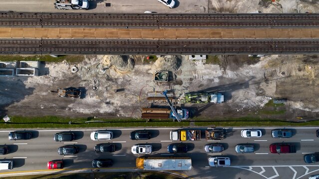 Aerial view of road construction repair site with heavy machinery, workers, next to moving car traffic and railway system on a sunny day