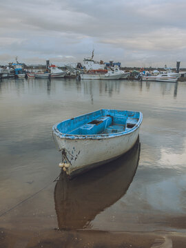 View of a bright blue and white rowboat resting on the shore reflecting in the still water of the harbor under a cloudy sky, Punta Umbr&iacute;a, Andalusia, Spain.