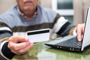An elderly man sits at a table with a laptop, holding a white bank card as he prepares to make an online payment, showing secure digital shopping.