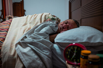 A senior man rests asleep in bed under soft blankets, with an alarm clock visible on the bedside table, depicting a calm nighttime home scene.