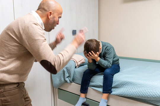 Angry adult man shouting at a young boy sitting on a bed, covering his face in distress. Indoor family conflict scene showing tension, discipline, and emotional stress. - Powered by Adobe