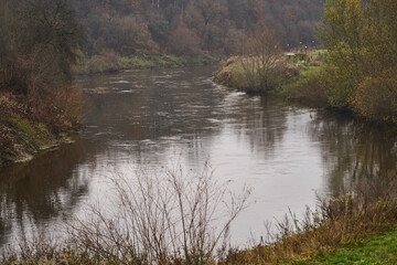 misty river bend with overcast sky, foggy autumn scene, bare trees and brown reeds framing...