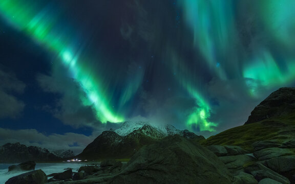 View of ethereal green aurora borealis dances above the tranquil, rocky shores and snow-capped mountains under a starlit sky, Henningsvaer, Nordland, Norway.