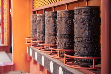 View of ornate, dark prayer wheels lined up against a vibrant orange wall, adorned with white dots, casting shadows in Diqing, Yunnan, China.