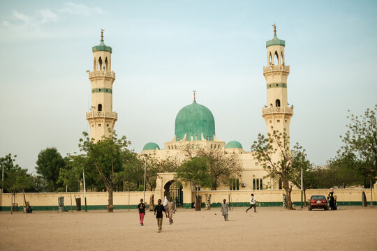 Kano, Nigeria - 26 May 2025: View of the Emir Palace Mosque, its pale cream walls and tall minarets contrasting with the vibrant turquoise dome and the expansive sandy foreground.