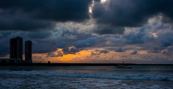 Lagos, Nigeria - 28 March 2025: View of Tarkwa Bay's serene waves meeting the shore as dark clouds loom above, a contrast to the distant buildings silhouetted against the orange sky.