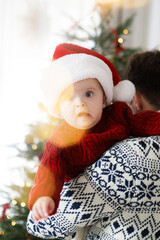 Happy father holding funny baby son in santa hat and knitted sweater against christmas tree lights. Baby's first christmas. Merry Christmas and Happy Holidays! Family holidays tradition