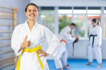Satisfied athlete girl stands in white kimono in gym during karate workout. Model gives thumbs up