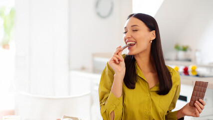 Young satisfied lady eating piece of black chocolate, holding tile in her hand, enjoying sweet...