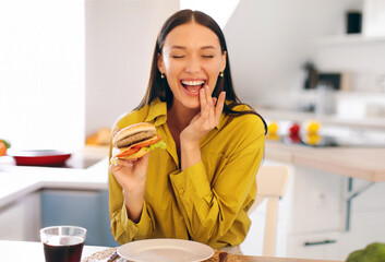 Gluttony. Satisfied lady enjoy eating hamburger, sitting in kitchen interior at home, copy space....