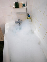 Bathtub filled with foam in a bright tiled bathroom. A faucet, shower head and shelf with bath products create a scene of relaxation and home self-care.
