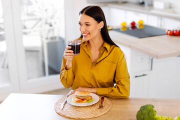 Happy European woman holding glass of cola while eating burger at home, sitting at table in kitchen interior and looking aside, copy space