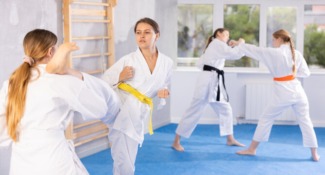 Focused young girl in white kimono practicing karate kicking techniques during sparring with female opponent in training room