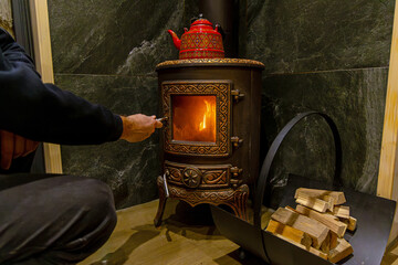 Man lighting, ornate wood stove with firewood. Person igniting a decorative cast iron wood-burning stove, with a red kettle on top and firewood beside it, indoors.