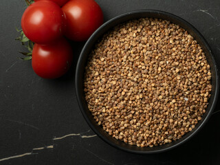 A bowl of raw buckwheat groats and cherry tomatoes as ingredients for a potava with a wooden spoon on a dark stone table