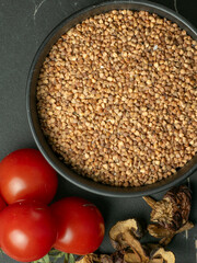 A bowl of raw buckwheat groats and cherry tomatoes as ingredients for a potava with a wooden spoon on a dark stone table