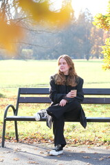 Girl is sitting on a bench in city autumn park with paper cup of coffee