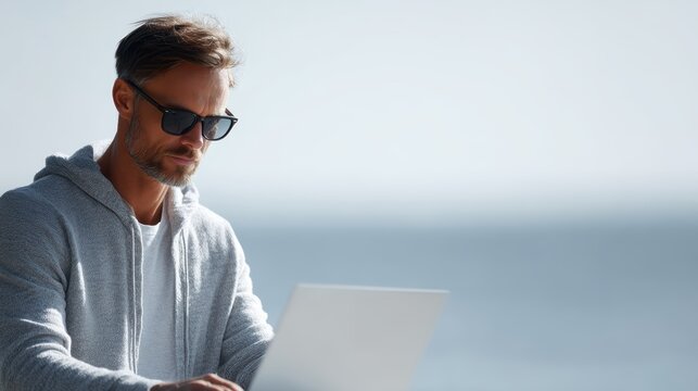 Pensive man freelancer in sunglasses typing on laptop by the sea, remote work vacation concept - Powered by Adobe