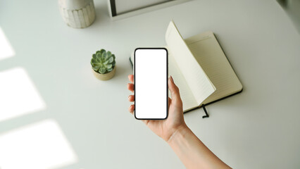 Female hand holding smartphone mockup over white desk, blank screen mobile phone template, open notebook diary background, minimalist top view workspace, for app, study, work, plan.