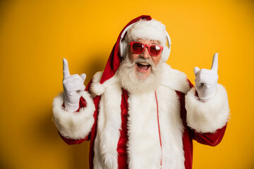 Santa Claus wearing headphones and sunglasses making rock and roll hand gesture isolated on yellow background